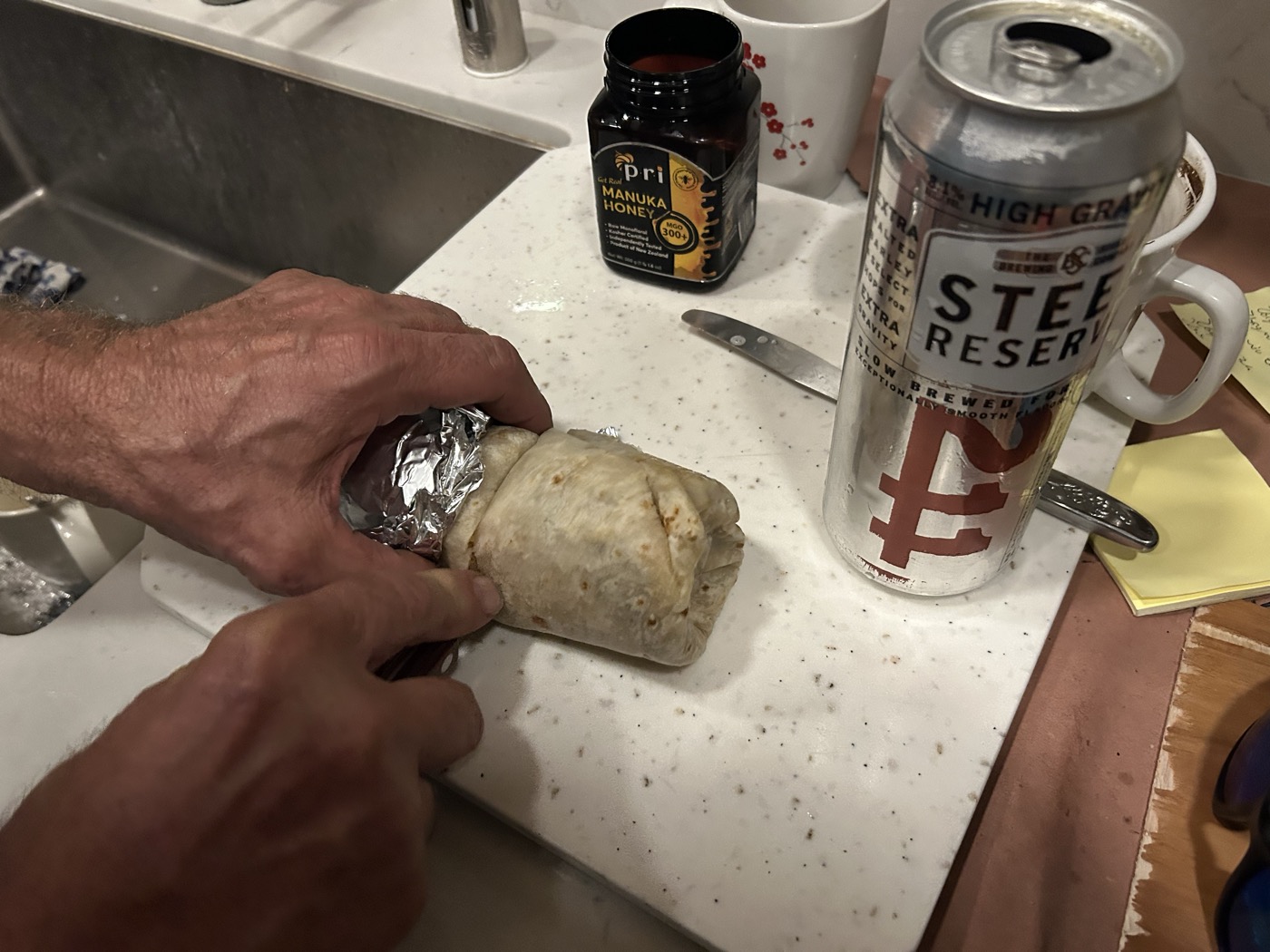 Blazej holding Los Pericos carne asada burrito, Steel Reserve High Gravity, Manuka Honey — McMorrow Rd kitchen — May 2026